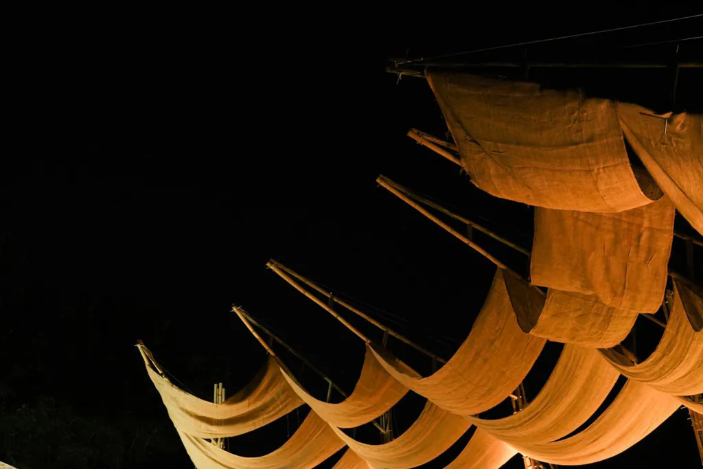 The Cantilevered Wave Pavilion at night: orange fabric panels swept up on bamboo arms, glowing against black