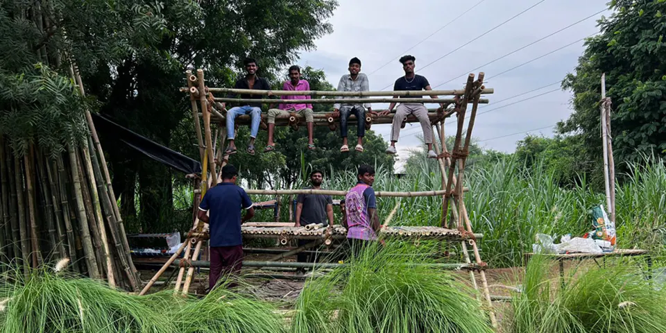 Four people sit on the upper deck of The X while others stand below, in a field of tall grass